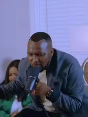 The cover image shows a man with a shaved head holding a microphone and singing out loud. He is wearing a dark grey blazer over a white shirt. There's a partially visible woman in the background on the left in a green shirt, and a neutral background setting.