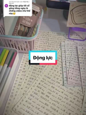 The image is a top-down shot of a desk covered with handwritten notes in a language that appears to be Chinese or a related Asian language. The notes are scattered across the surface, along with colorful markers, a pink and white basket, and a box of what seems to be coffee-flavored soft bread. The focal point is a rectangular text box in teal and white that says "Động lực" (which translates to "Motivation"). The overall composition suggests a study or work environment, with a focus on language learning.