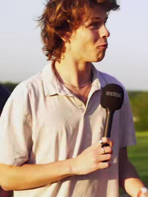 A young man with brown curly hair is holding a black microphone with a Rode logo. He is wearing a light-colored button-down shirt and a silver necklace. Some greenery and sky are slightly visible in the background.