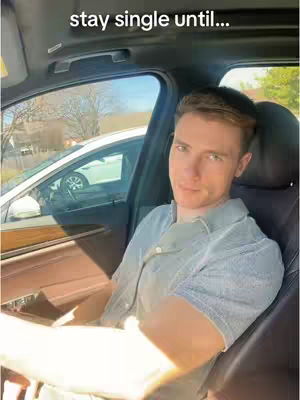 A man is sitting in the driver's seat of a car, looking at the camera with a neutral expression. The car's interior and the obscured exterior environment are visible, suggesting an indoor background with sunlight filtering through the windows.