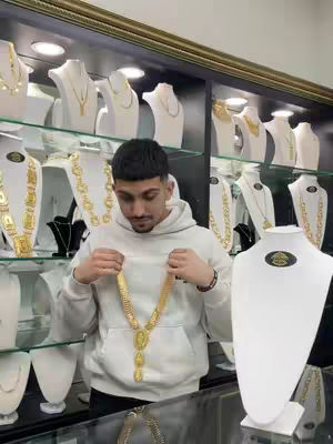 A man in a white hoodie holds up a gold necklace in a jewelry store. Gold necklaces are displayed on white busts behind him.