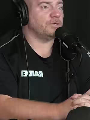 A man wearing a black shirt and headphones, holding a microphone. The background is dark. The shirt has white text, and the man is speaking.