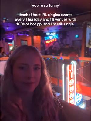 A selfie-style image taken in a bar with the creator in the foreground. The background is blurred, with neon signs and other indistinct figures. The creator, a blonde woman, is centered and looking towards the camera. Text is overlaid at the top.