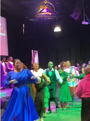 A group of people dancing on a stage. The lighting is dim with a couple of spotlights and a church banner in the background and logo. The foreground has people dressed in colorful outfits, with a lady wearing a blue dress in the front.
