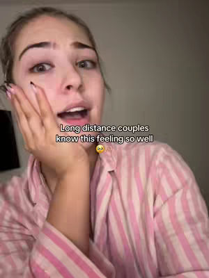 A woman with light skin and blonde hair is shown in a close-up shot, looking directly at the camera with a slightly open mouth. She has a hand near her face. The background is blurred, but appears to be a dimly lit room.