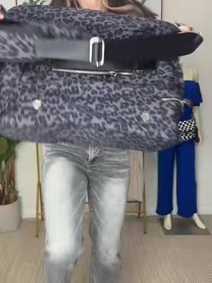 A woman is standing and holding a large bag with a leopard print design in front of her. She wears grey denim jeans and the background includes a clothes rack with a dress and a mannequin wearing a blue outfit.