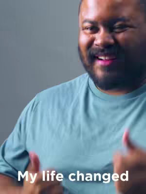 A medium shot shows a man with a dark complexion and a full beard, smiling broadly and giving a thumbs-up with both hands. He's wearing a light blue t-shirt. The background is a neutral grey. The words 'My life changed' are overlaid at the bottom of the image.