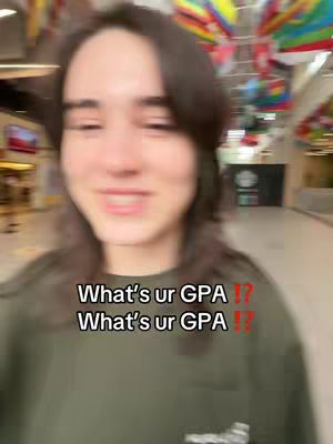 A selfie-style shot featuring a person looking directly at the camera, with text overlayed. The individual has dark hair and is wearing a dark green shirt. The background is slightly blurred but appears to be a public space with a variety of colors and structures.