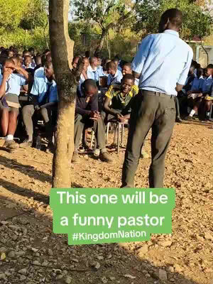 The image shows a group of people in what appears to be a casual outdoor setting, possibly a school or community gathering. A large tree trunk is in the foreground on the left, a man is standing in the center, and a group of people are sitting on the ground. There is a green banner overlayed at the bottom of the image with text.