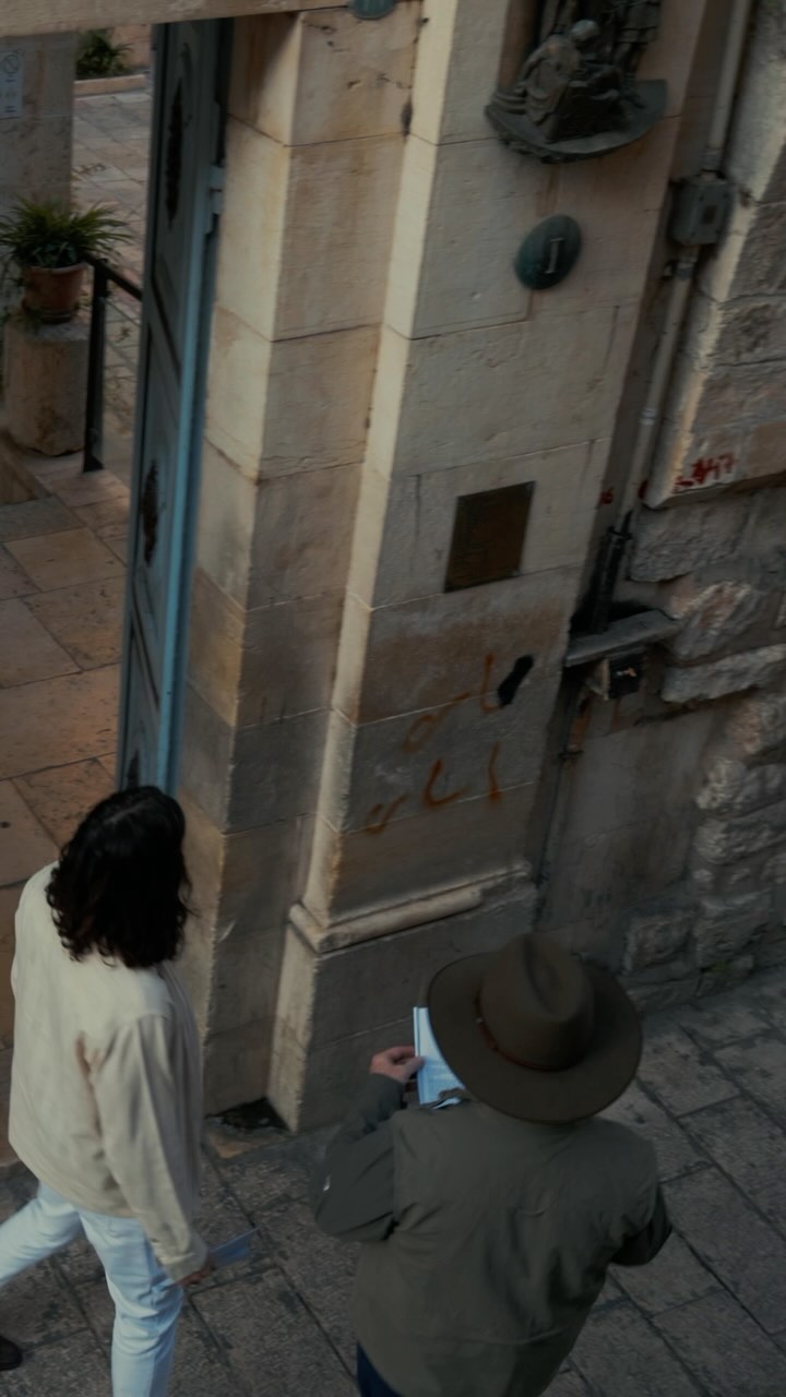 An overhead shot shows two people walking down a stone-paved street alleyway in an old city. One person is in front with their back to the camera, wearing a cream jacket, white pants, and black hair. The other person is behind holding an open booklet, wearing a hat and a green jacket. The alleyway walls are made of stone with a doorway to the left side.