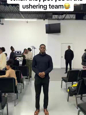 A man in a dark navy sweater and black pants stands in the center of a brightly lit room, likely a church. Several other people are in the background, some seated and others standing. Text overlays the image.
