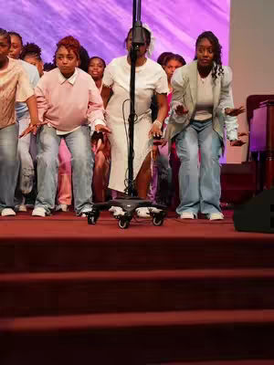 A group of people, mostly women, are on stage in a church setting. There is a microphone stand in the center. The stage is lit with a purple background.