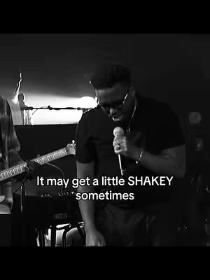 A black and white cover image featuring a man in a dark setting, likely a stage, holding a microphone. Text overlayed on the image reads "It may get a little SHAKEY sometimes". The man is wearing glasses and a dark shirt, with what appears to be a band or musicians behind him. The lighting is dim, and the overall composition is focused on the man with the microphone.