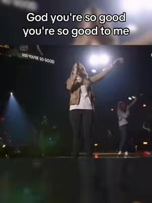 A woman singing on stage with arms raised, with additional people visible in the background, with on-screen text overlay: God you're so good, you're so good to me.