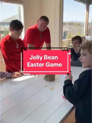 A group of people, including a man and several children, are gathered around a white table. The focus is on colorful jelly beans scattered on the table and a red text overlay that reads 'Jelly Bean Easter Game'. The scene is indoors with a window in the background.