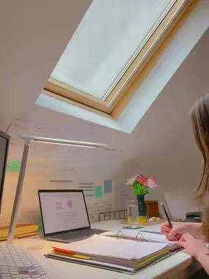 A room with a slanted ceiling and a large skylight. A desk is set up with a laptop, a blank notebook, and some stationary. The room is lit with a desk lamp and natural light coming from a skylight. A person is seated at the desk and is visible from the shoulders down and is writing.
