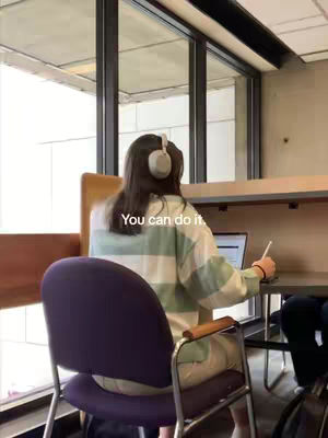 A person is sitting at a desk, viewed from behind. The individual is wearing a white and green striped shirt and headphones. A laptop is open on the desk. The text reads 'You can do it.'