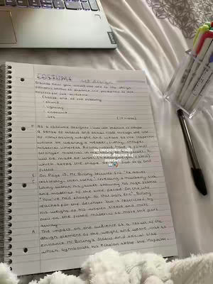 A spiral-bound notebook with handwritten notes on a white surface, possibly a bed. A laptop and a pen on a clear holder filled with colorful markers are also on the surface. The image is lit with natural light.