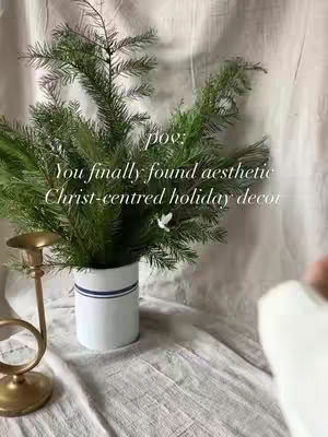 An aesthetic shot of a white vase with a blue stripe around its center and green foliage inside, sits on a wrinkled cream-colored cloth. A golden candlestick is present on the left side, and a neutral background.