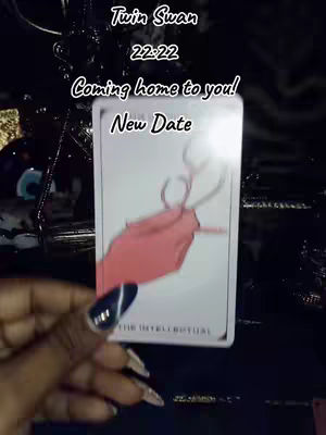A close-up shot shows a hand holding a tarot card against a dark surface. The card features a drawing of a pair of antlers connected by a line, colored in reddish-pink, on a white background. The hand has long, manicured nails. Above the card, text reads 'Twin Swan 22:22.' Additional text on the card says 'Coming home to you!' and 'New Date.' The background appears to be a dark, possibly shiny material, with subtle reflections and shadows.