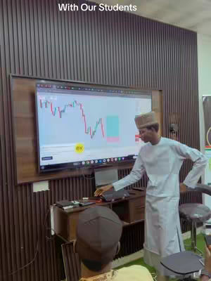 A man wearing a white outfit and a cap is standing in front of a large screen displaying a cryptocurrency chart. The setting appears to be a room with wooden paneling on the walls, and an audience of individuals sitting in front of the screen, listening to the speaker. The image is well-lit, with the focus on the screen.