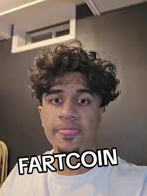 A selfie-style shot featuring a young man with dark, curly hair, positioned against a simple indoor backdrop. The man is looking towards the camera with a slightly playful expression, his tongue sticking out. Below his face, the text "FARTCOIN" is boldly displayed in a white, outlined font. The color palette is neutral, with the man wearing a basic white t-shirt.