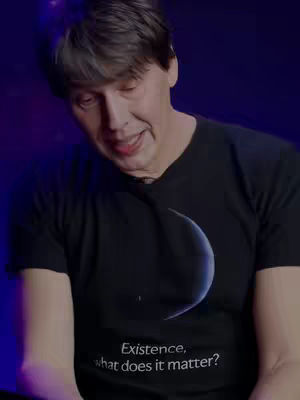 A medium shot features a man with short, dark hair and a slightly aged appearance, wearing a black t-shirt. The lighting suggests a dim, indoors setting, possibly a studio, with a blurred blue background adding a subtle contrast to the scene. The man appears to be addressing the camera, his expression open and engaged, and a graphic on his shirt provides an intriguing visual element.