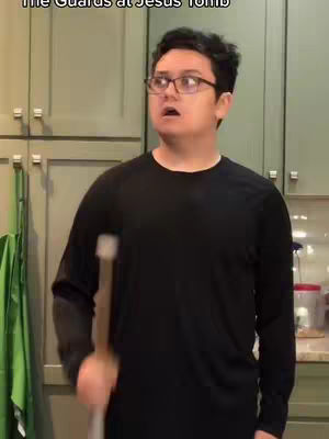 A man with glasses looks surprised. He is holding a stick and wearing a black long-sleeved shirt, standing in front of some kitchen cabinets.