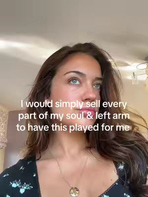 A selfie-style shot featuring a young woman with long brown hair, blue eyes, and fair skin. She is looking towards the side with a thoughtful expression. She wears a black top with a floral design and a delicate necklace. Text overlayed indicates a desire for someone to play a specific song.