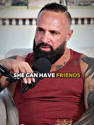 A man with a beard and tattoos is holding a microphone with the text overlayed. The man is wearing a red tank top and appears to be in a casual setting.