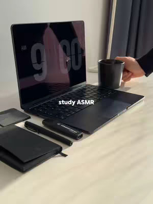 A minimalist setting featuring a black laptop open on a white desk, with a notebook, pens, and a coffee cup. The screen displays the time. The composition is clean and organized, lit by natural light.
