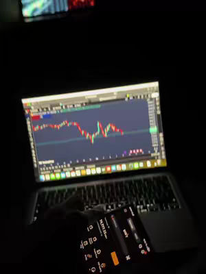 A dimly lit scene featuring a laptop displaying a stock chart and a smartphone displaying financial data. The laptop screen shows red and green candlestick patterns, indicating market activity. The person's hand is holding the phone which shows numbers. The background is completely dark.