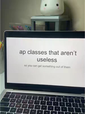 A laptop is open, displaying the words "ap classes that aren't useless", and below it, "so you can get something out of them". The laptop is on a desk with various other items visible, including a small decorative item and some pens.