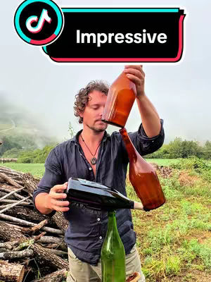 A man balances three bottles in a natural outdoor setting. The foreground includes a pile of logs and green grass, and the background is filled with trees and mist. The focus is on the man, who is holding the bottles with concentration, and the bottles themselves are brown, green and clear
