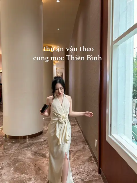 A woman in a cream-colored dress is walking in a well-lit hallway. The dress has a halter neck and a slit, and she's holding a black phone. Text overlays the image about dressing according to the astrological sign.