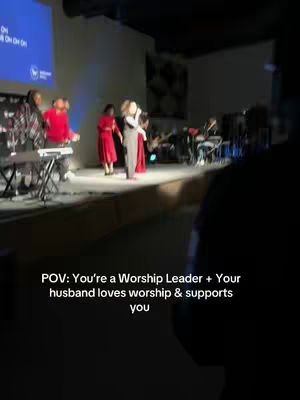 The image captures a live performance on a stage, indoors. In the foreground, a dark subject obscures a portion of the scene. On the stage, a group of people are present, with a central figure holding a microphone, likely a worship leader. Other figures, including those playing instruments, are visible to the left and right. A blue screen is in the background. The lighting is dim, which gives the image a performance setting.