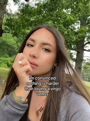 A young woman with long brown hair, wearing a gray hoodie, is the central focus. She's in a natural outdoor setting with trees in the background. The woman has a contemplative expression, resting her chin on her hand, with a small ring visible. Text overlayed on the image reads, 'im convinced nothing is harder than loving a virgo venus.'