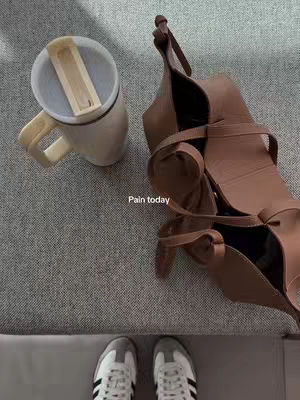 An overhead shot shows a casual scene: a half-empty mug with a closed lid, a brown handbag, and a pair of sneakers against a gray textural surface. The composition suggests a workspace or everyday setting, with a focus on personal items.