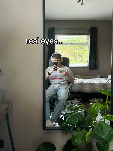 A mirror selfie of a man sitting in a chair. The man is wearing headphones, a t-shirt, and jeans. He's holding a phone in front of his face with his left hand and a mug in his right hand. The video reflection shows a room with a bed, a window with curtains, and green outdoor plants with natural lighting.