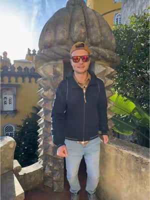 A man wearing sunglasses and a cap stands in front of a textured stone structure. The background shows a building and greenery under a bright sky.