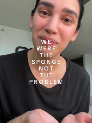 A close-up shot of a woman's face looking towards the camera. Text overlaid on the image reads: 'WE WERE THE SPONGE NOT THE PROBLEM'. The woman has dark hair and is wearing a black top, making the text stand out. The background is softly lit and blurred.
