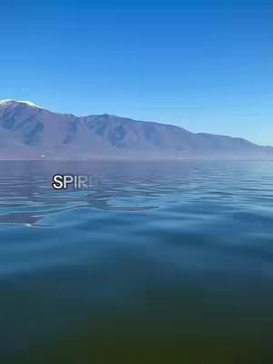 A serene landscape featuring a body of water in the foreground, leading towards a distant mountain range under a clear blue sky. The water reflects the sky, and the overall composition creates a sense of calm.