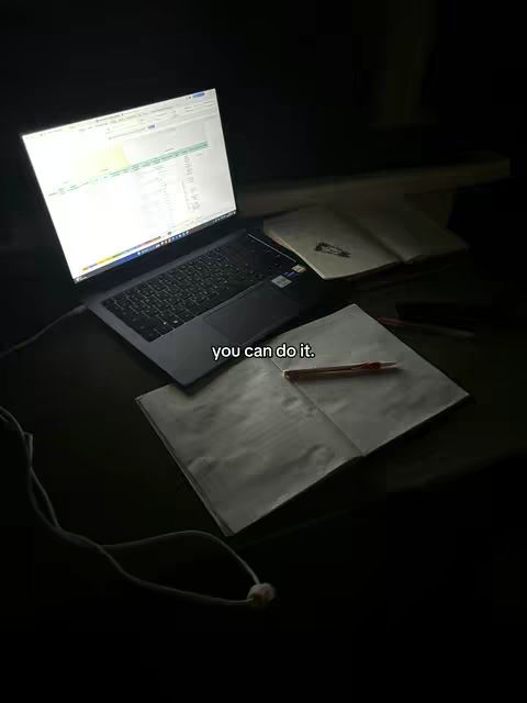 A dimly lit scene captures a laptop with a brightly illuminated screen displaying what appears to be a spreadsheet, casting a glow onto the surrounding area. To the right of the laptop, an open notebook and a pen lie on a desk. The image is dark, emphasizing the screen's light and the objects on the surface.