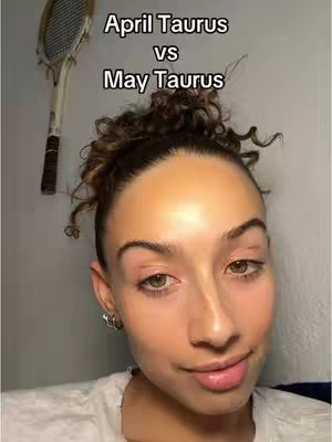 A selfie-style image with a light-skinned woman facing the camera. She has dark curly hair pulled back, with some strands framing her face. The background looks to be a plain wall with a tennis racket hanging on it. A text overlay at the top reads "April Taurus vs May Taurus". The woman is wearing a white shirt and has minimal makeup.