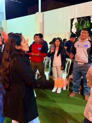 A group of people is gathered outdoors, likely at an event or gathering. In the foreground, a woman in a black coat gestures with her hands in the center, looking towards the right. She is the main focus of the image and appears to be speaking or singing. Others are in the background, with a man in a red shirt, various people in casual attire, and white plastic chairs visible.