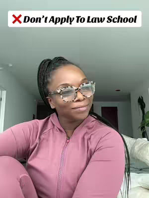 A woman with dark braids and large framed glasses sits in front of a blurred background. She wears a pink zip-up jacket and is angled slightly towards the right.