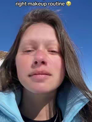 A selfie-style shot of a young woman with long, brown hair against a bright blue sky. The woman is looking slightly down with a neutral expression. She's wearing a light blue jacket. The text "right makeup routine 🥺" is overlayed at the top of the video.