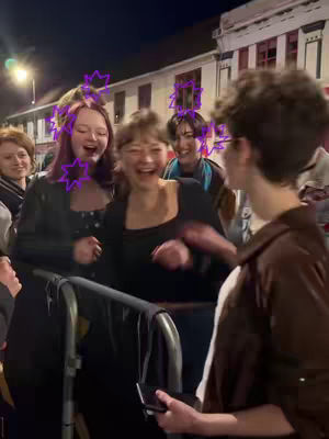 A group of young people are gathered outdoors at night in front of a building. They are laughing and appear to be enjoying a social gathering. The scene is lit by streetlights, creating a warm tone. The people are dressed in casual wear.