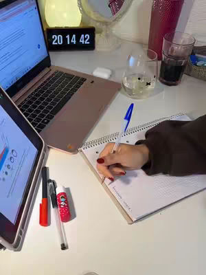 A desk setup featuring a laptop, tablet, and notebook with a hand writing. The setting is well-lit with soft lighting, and various stationery items are visible. A clock displays the time.