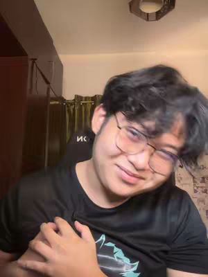 A young man with glasses smiles while looking at the camera. He is wearing a black t-shirt and his dark hair is slightly messy. The background is a blurred room interior.